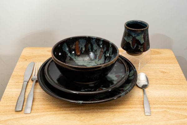the photograph shows a clay coyote tableware setting. it is 4 pieces of clay coyote pottery, a dinner plate, a sandwich plate, a soup and chili bowl and a yunomi cup. in the photograph the tableware setting pieces are all glazed in midnight garden. there is a fork and a knife on the left hand side of the stack of 2 plates and bowl. on the right side of the stack there is a spoon. in the upper right there is the yunomi on a small clear plastic stand. everything is resting on a small light colored wooden table. the background is a white wall. the photograph is lit with white light.