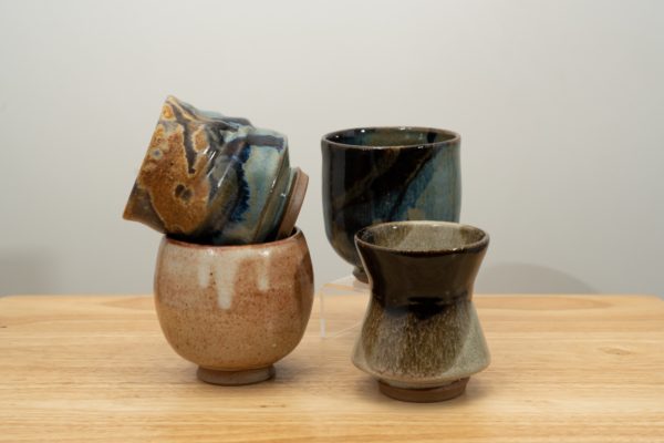 the photograph shows 4 clay coyote yunomi wine and tea cups resting on a small light colored wooden table. the 4 cups all have different glazes. starting from the left, they go: a shino glazed cup with a joes blue glazed up resting diagonally on top of it. to the right and slightly closer to the camera is a mint chip glazed yunomi. behind the mint chip yunomi is one glazed in zappa. the zappa yunomi is resting on a small clear plastic stand, allowing most of it to be visible even though it is placed behind the mint glazed one. the background is a white wall.