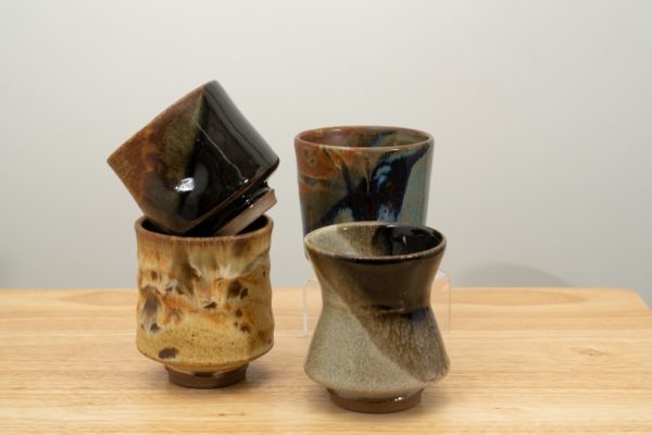 the photograph shows 4 clay coyote yunomi wine and tea cups resting on a small light colored wooden table. two of the cups are stacked on on the other, the base of the two stack is a feather glazed yunomi, with a mocha swirl cut resting diagonally inside the feather cup. to the right of these two and slightly closer to the camera is a mint chip glazed yunomi. behind the mint chip yunomi is the last cup, glazed in joes blue, and resting on a small clear plastic stand, making most of it visible, even though it is behind the mint chip cup. the background is a white wall. the photograph is lit with white light.