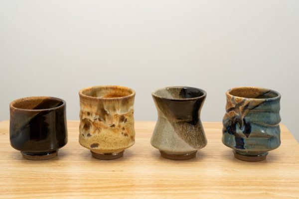 the photograph shows 4 clay coyote yunomi wine and tea cups in a line. from left to right the glazes are mocha swirl, feather, mint chip and joes blue. all the cups are resting on a small light colored wooden table. the background is a white wall. the photograph is lit with white light.