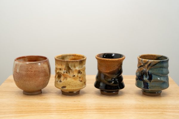 the photograph shows 4 clay coyote yunomi wine and tea cups in a line. from left to right the glazes are shino, feather, mocha swirl and joes blue. all the cups are resting on a small light colored wooden table. the background is a white wall. the photograph is lit with white light.