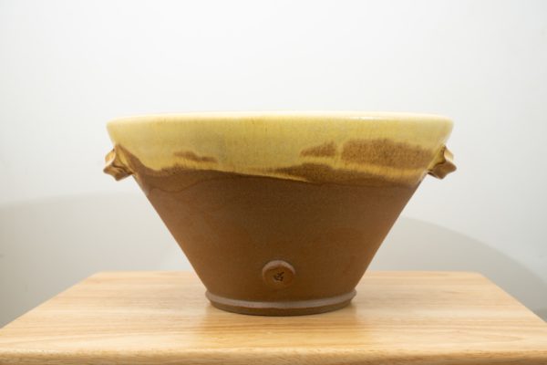 the photograph shows a clay coyote original cassole resting flat on a small light colored wooden table. the cassole has a small button facing the camera with the clay coyote coyote stamped into it. the cassole has yellow salt glaze on the upper outside lip (and all of the inside), just past the two handles on opposite sides. the background is a plain white wall. the photograph is lit with white light.
