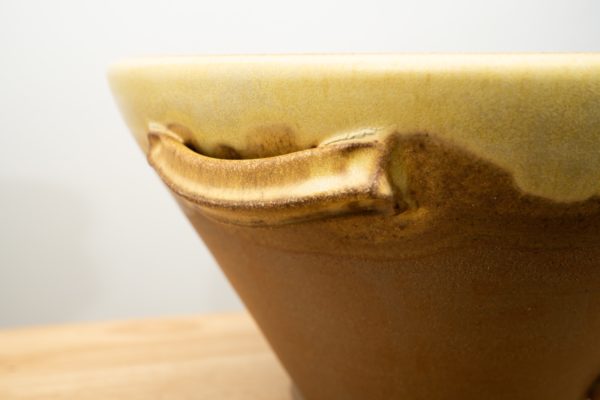 the photograph shows a close up of the handle of a clay coyote original cassole. the handle and upper edge of the cassole are glazed in yellow salt. below the handle the cassole is not glazed, showing the reddish brown of the natural clay. only the upper edge, handle and a small part of the cassole are visible due to the closeness of the photograph. the background is a plain white wall. near the bottom left corner the table that the cassole is resting on is slightly visible. it is light colored wood, and is out of focus.