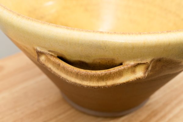 the photograph shows a close up, high angle view of one of the two handles on a clay coyote original cassole. the cassole is glazed in yellow salt. the handle and the upper edge of the cassole are glazed, while the outside bottom of the cassole is unglazed and shows the color of the clay (reddish brown). the table the cassole is resting on is slightly visible in the bottom left and right corners of the photograph. it is a small light colored wooden table. on the upper edge of the photograph the yellow salt glaze on the inside of the cassole is visible.