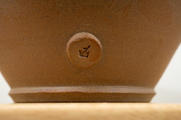 the photograph shows the base of a original clay coyote cassole. the "front" of the cassole is pointed towards the camera, with a close up of the small button near the base of the cassole. the button has the clay coyote coyote makers mark on it. only the bottom base of the cassole is visible in this photograph. the background is a white wall out of focus. this part of the cassole is unglazed and shows the natural reddish brown color of the clay.