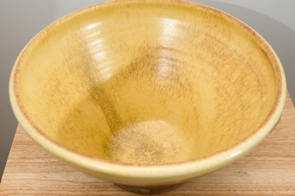 the photograph shows a high angle view of the inside of a clay coyote original cassole. the cassole is glazed in yellow salt. the entirety of the inside of the bowl is glazed. the cassole is resting on a small light colored wooden table. the table is sitting in front of a blank white wall. the photograph is lit with white light.