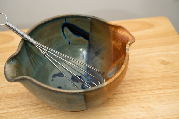the photograph shows a high angle view of a clay coyote mixing bowl with whisk glazed in joes blue. the wire metal whisk is inside the bowl. the whisks handle is pointed roughly towards the upper left corner of the photograph. the angle of the photograph allows the viewer to see the joes blue pattern on the inside of the mixing bowl. the mixing bowl is resting on a small light colored wooden table.