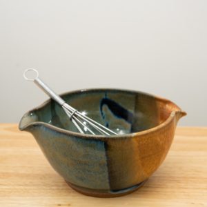 the photograph shows a clay coyote mixing bowl glazed in joes blue. the mixing bowl has a wire whisk resting inside of it. the wire whisk handle is pointed roughly towards the upper left corner. the mixing bowl is resting on a small light colored wooden table, in front of a white wall. the photograph is lit with white light.