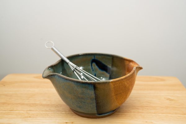 the photograph shows a clay coyote mixing bowl glazed in joes blue. the mixing bowl has a wire whisk resting inside of it. the wire whisk handle is pointed roughly towards the upper left corner. the mixing bowl is resting on a small light colored wooden table, in front of a white wall. the photograph is lit with white light.