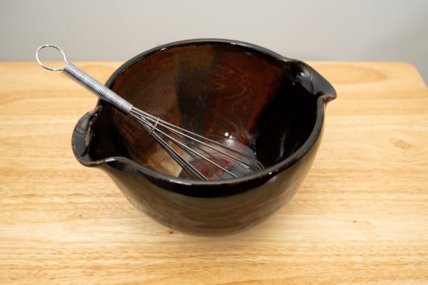 the photograph shows a high angle view of a clay coyote mixing bowl with whisk glazed in merlot. the wire metal whisk is inside the bowl. the whisks handle is pointed roughly towards the upper left corner of the photograph. the angle of the photograph allows the viewer to see the merlot pattern on the inside of the mixing bowl. the mixing bowl is resting on a small light colored wooden table.