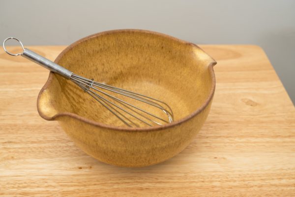 the photograph shows a high angle view of a clay coyote mixing bowl with whisk glazed in yellow salt. the wire metal whisk is inside the bowl. the whisks handle is pointed roughly towards the upper left corner of the photograph. the angle of the photograph allows the viewer to see the inside of the mixing bowl. the mixing bowl is resting on a small light colored wooden table.
