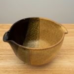 the photograph shows a high angle view of a clay coyote mixing bowl glazed in tiger. the mixing bowl is resting on a small light colored wooden table. the background is a plain white wall.