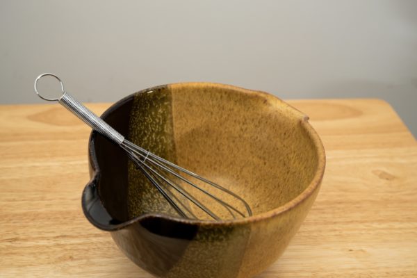 the photograph shows a high angle view of a clay coyote mixing bowl with whisk glazed in tiger. the wire metal whisk is inside the bowl. the whisks handle is pointed roughly towards the upper left corner of the photograph. the angle of the photograph allows the viewer to see the tiger pattern on the inside of the mixing bowl. the mixing bowl is resting on a small light colored wooden table.