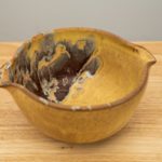 the photograph shows a high angle view of a clay coyote mixing bowl glazed in tequila sunrise. the angle allows the viewer to see the tequila sunrise pattern of the glaze inside the mixing bowl. the mixing bowl is resting on a small light colored wooden table. the background is a plain white wall.