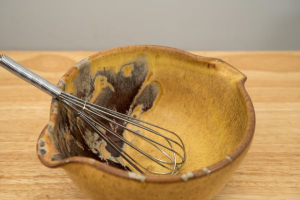 the photograph shows a high angle view of a clay coyote mixing bowl with whisk glazed in tequila sunrise. the wire metal whisk is inside the bowl. the whisks handle is pointed roughly towards the upper left corner of the photograph. the angle of the photograph allows the viewer to see the tequila sunrise pattern on the inside of the mixing bowl. the mixing bowl is resting on a small light colored wooden table.