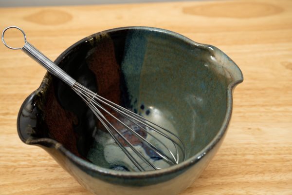 the photograph shows a high angle view of a clay coyote mixing bowl with whisk glazed in zappa. the wire metal whisk is inside the bowl. the whisks handle is pointed roughly towards the upper left corner of the photograph. the angle of the photograph allows the viewer to see the zappa pattern on the inside of the mixing bowl. the mixing bowl is resting on a small light colored wooden table.