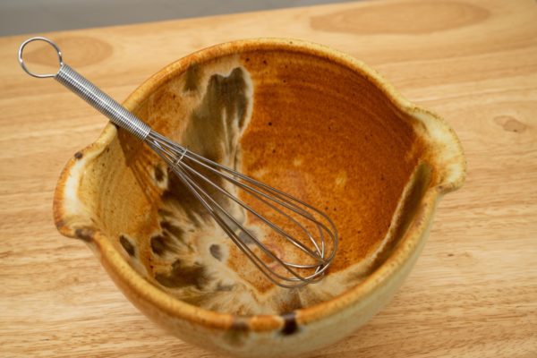 the photograph shows a high angle view of a clay coyote mixing bowl with whisk glazed in feather. the wire metal whisk is inside the bowl. the whisks handle is pointed roughly towards the upper left corner of the photograph. the angle of the photograph allows the viewer to see the feather pattern on the inside of the mixing bowl. the mixing bowl is resting on a small light colored wooden table.