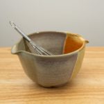 the photograph shows a clay coyote mixing bowl with whisk glazed in mint berry with a wire metal whisk inside. the whisks handle is pointed roughly towards the upper left corner of the photograph. the mixing bowl is resting on a small light colored wooden table. the background is a plain white wall.