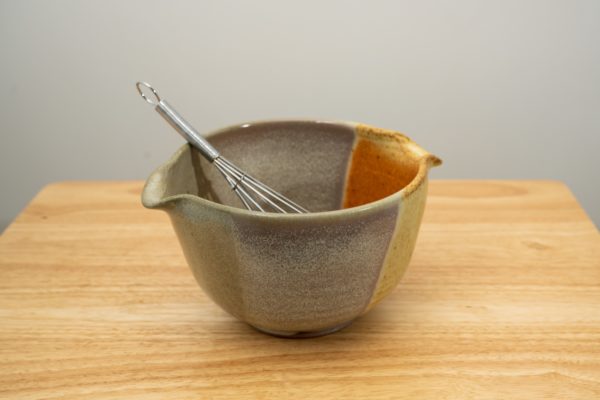 the photograph shows a clay coyote mixing bowl with whisk glazed in mint berry with a wire metal whisk inside. the whisks handle is pointed roughly towards the upper left corner of the photograph. the mixing bowl is resting on a small light colored wooden table. the background is a plain white wall.