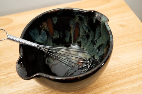 the photograph shows a clay coyote mixing bowl glazed in midnight garden. the mixing bowl has a wire whisk resting inside of it. the wire whisk handle is pointed roughly towards the upper left corner. the mixing bowl is resting on a small light colored wooden table, in front of a white wall. the photograph is lit with white light.