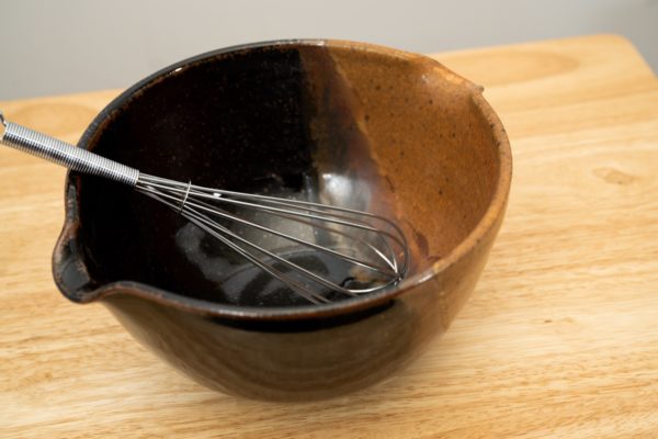 the photograph shows a clay coyote mixing bowl glazed in mocha swirl. the mixing bowl has a wire whisk resting inside of it. the wire whisk handle is pointed roughly towards the upper left corner. the mixing bowl is resting on a small light colored wooden table, in front of a white wall. the photograph is lit with white light.