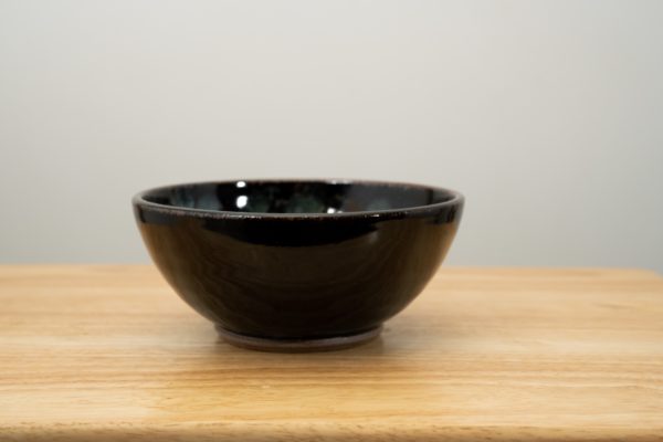 the photograph shows a clay coyote soup and chili bowl glazed in midnight garden. the bowl is resting on a light colored wooden surface. the glaze pattern for midnight garden is on the inside of the bowl, and is barely visible on the back rim. the side of the bowl facing the camera is plain black. the photograph is lit with white light. the background is a white wall.