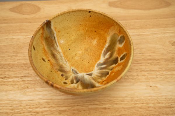 the photograph shows a high angle view of a clay coyote soup and chili bowl glazed in feather. the angle of the viewpoint allows the viewer to see inside the bowl, and to see the feather pattern it contains. the bowl is resting on a light colored wooden surface. the photograph is well lit with white light.
