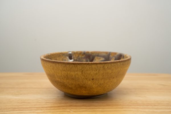 the photograph shows a clay coyote soup and chili bowl glazed in tequila sunrise resting on a light colored wooden surface. the photograph angle is just a bit above the rim of the bowl. the outside of the bowl is yellow. the background is a white wall.