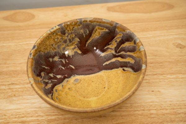 the photograph shows a high angle view of a clay coyote soup and chili bowl glazed in tequila sunrise. the angle of the photograph allows the viewer to see into the bowl and see the color pattern of the tequila sunrise glaze on this inside of the bowl. the bowl is resting on a light colored wooden surface.