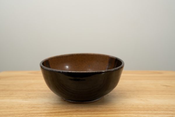 the photograph shows a clay coyote soup and chili bowl glazed in mocha swirl resting on a light colored wooden table. the background is a white wall. the photograph is well lit with white light.