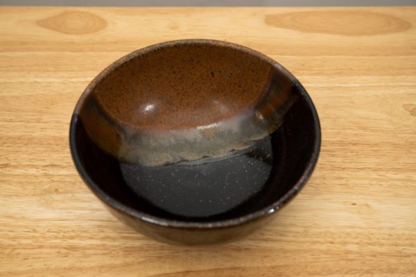 the photograph shows a high angle view of a clay coyote soup and chili bowl glazed in mocha swirl. the angle of the view allows the viewer to see the pattern of the mocha swirl in the bowl. the bowl is resting on a light colored wooden surface. the photograph is lit with white light.