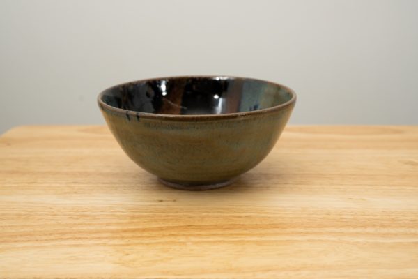 the photograph shows a clay coyote soup and chili bowl glazed in zappa. the bowl is resting flat on a small light colored wooden table. the background is a white wall. the photograph is well lit with white light.