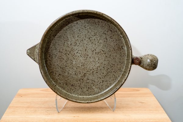 the photograph shows a clay coyote flameware cazuela resting on a clear plastic stand. the stand allows the cazuela to stand almost vertically, this allows the viewer a full look at the cooking surface. the bulbous handle is pointed to the right, and the small secondary triangular handle is pointed to the right. the cazuela is glazed in coyote grey (grey with darker speckling through out). the cazuela is resting on a small light colored wooden table. the background is a white wall.