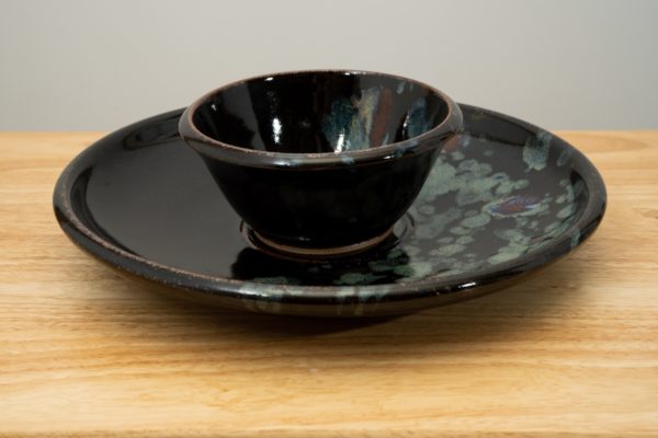the photograph shows a high angle view of a clay coyote chip and dip set glazed in midnight garden resting flat on a small light colored wooden table. the chip and dip set consists of a large platter and a smaller dip bowl that fits in the center of the platter. the bowl is resting in the small indented area in the center of the platter as it was designed to. the background is a white wall. the photograph is lit with white light.