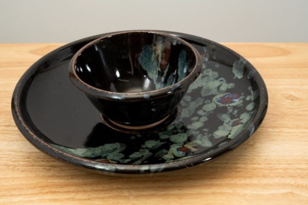 the photograph shows a clay coyote chip and dip set glazed in midnight garden resting flat on a small light colored wooden table. the chip and dip set consists of a large platter and a smaller dip bowl that fits in the center of the platter. the bowl is resting in the small indented area in the center of the platter as it was designed to. the background is a white wall. the photograph is lit with white light.