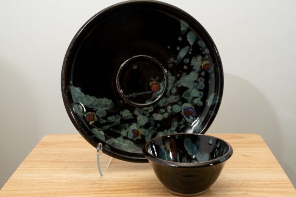 the photograph shows two clay coyote pieces on a small light colored wooden table. the two pieces are the clay coyote chip and dip set glazed in midnight garden. the large platter is upright resting on a clear plastic stand, to allow a good view of the midnight garden glaze pattern. to the front and slight right of the patter and stand is the dip bowl. it is sitting flat on its base on the small wooden table. the background is a white wall. the photograph is lit with white light.