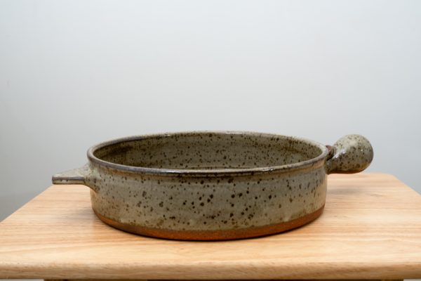 the photograph shows a clay coyote flameware cazuela resting on a small light colored wooden table. the cazuela is glazed in coyote grey (grey with darker speckling through out). the cazuelas main bulbous handle is pointed to the right. the secondary triangular handle is pointed to the right. the cazuela is laying flat on the table. the background is a plain white wall. the photograph is lit with white light.