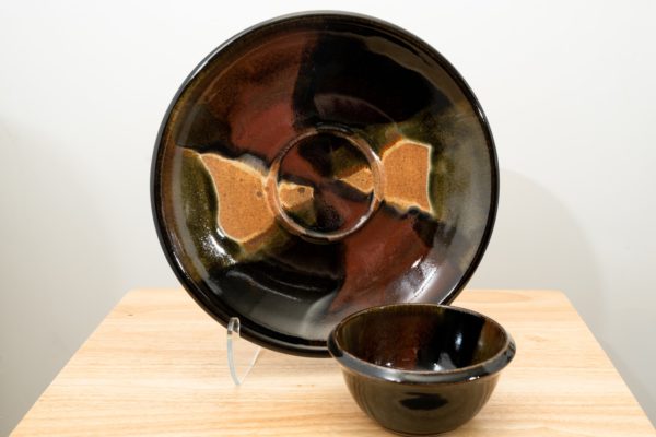 the photograph shows two clay coyote pieces on a small light colored wooden table. the two pieces are the clay coyote chip and dip set glazed in merlot. the large platter is upright resting on a clear plastic stand, to allow a good view of the merlot glaze pattern. to the front and slight right of the patter and stand is the dip bowl. it is sitting flat on its base on the small wooden table. the background is a white wall. the photograph is lit with white light.
