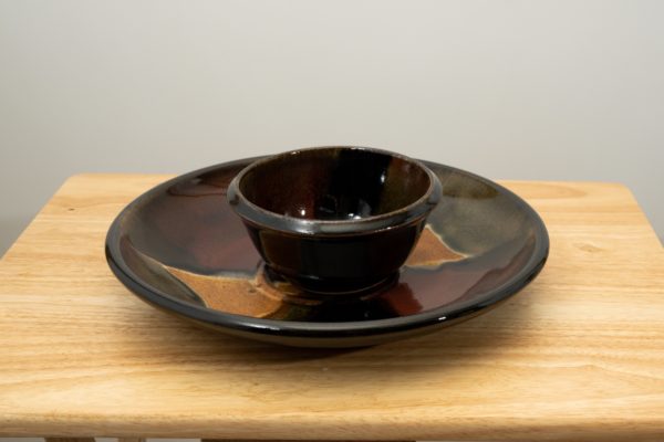 the photograph shows a clay coyote chip and dip set glazed in merlot resting flat on a small light colored wooden table. the chip and dip set consists of a large platter and a smaller dip bowl that fits in the center of the platter. the bowl is resting in the small indented area in the center of the platter as it was designed to. the background is a white wall. the photograph is lit with white light.