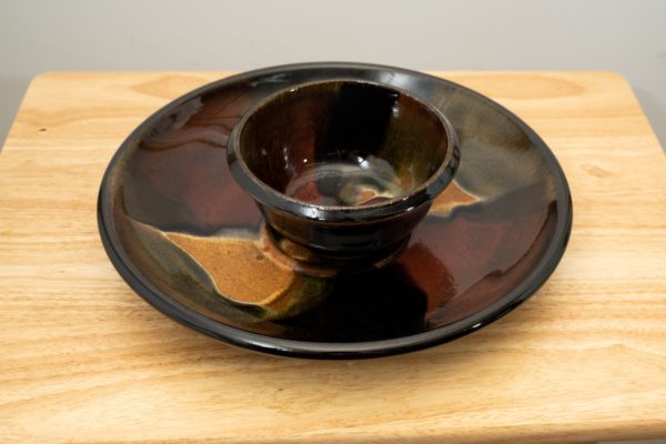 the photograph shows a high angle view of a clay coyote chip and dip set glazed in merlot resting flat on a small light colored wooden table. the chip and dip set consists of a large platter and a smaller dip bowl that fits in the center of the platter. the bowl is resting in the small indented area in the center of the platter as it was designed to. the background is a white wall. the photograph is lit with white light.