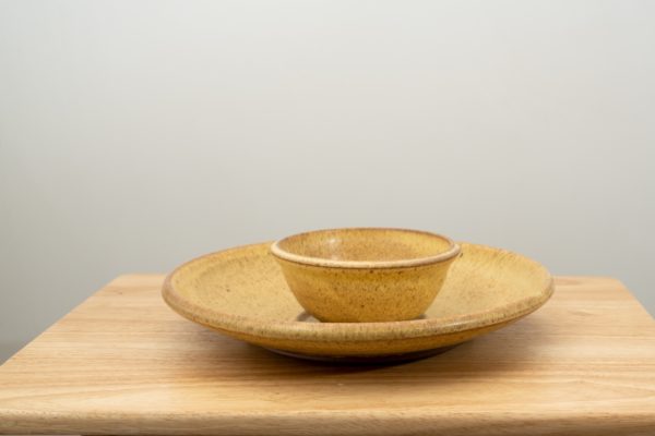 the photograph shows a clay coyote chip and dip set glazed in yellow salt resting flat on a small light colored wooden table. the chip and dip set consists of a large platter and a smaller dip bowl that fits in the center of the platter. the bowl is resting in the small indented area in the center of the platter as it was designed to. the background is a white wall. the photograph is lit with white light.