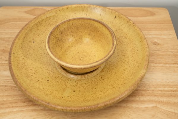 the photograph shows a high angle view of a clay coyote chip and dip set glazed in yellow salt resting flat on a small light colored wooden table. the chip and dip set consists of a large platter and a smaller dip bowl that fits in the center of the platter. the bowl is resting in the small indented area in the center of the platter as it was designed to. the background is a white wall. the photograph is lit with white light.