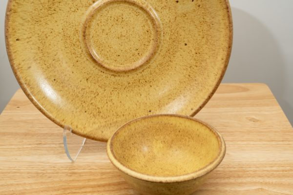 the photograph shows two clay coyote pieces on a small light colored wooden table. the two pieces are the clay coyote chip and dip set glazed in yellow salt. the large platter is upright resting on a clear plastic stand, to allow a good view of the yellow salt glaze pattern. to the front and slight right of the patter and stand is the dip bowl. it is sitting flat on its base on the small wooden table. the background is a white wall. the photograph is lit with white light.