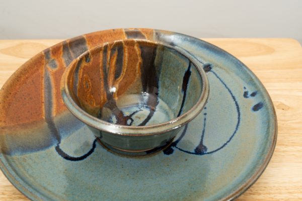 the photograph shows a high angle view of a clay coyote chip and dip set glazed in joes blue resting flat on a small light colored wooden table. the chip and dip set consists of a large platter and a smaller dip bowl that fits in the center of the platter. the bowl is resting in the small indented area in the center of the platter as it was designed to. the background is a white wall. the photograph is lit with white light.