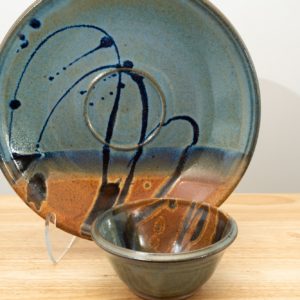 the photograph shows two clay coyote pieces on a small light colored wooden table. the two pieces are the clay coyote chip and dip set glazed in joes blue. the large platter is upright resting on a clear plastic stand, to allow a good view of the joes blue glaze pattern. to the front and slight right of the patter and stand is the dip bowl. it is sitting flat on its base on the small wooden table. the background is a white wall. the photograph is lit with white light.