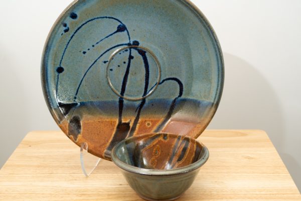 the photograph shows two clay coyote pieces on a small light colored wooden table. the two pieces are the clay coyote chip and dip set glazed in joes blue. the large platter is upright resting on a clear plastic stand, to allow a good view of the joes blue glaze pattern. to the front and slight right of the patter and stand is the dip bowl. it is sitting flat on its base on the small wooden table. the background is a white wall. the photograph is lit with white light.