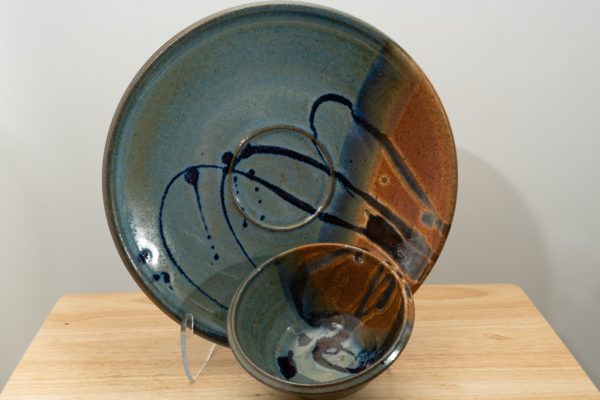 the photograph shows two clay coyote pieces on a small light colored wooden table. the two pieces are the clay coyote chip and dip set glazed in joes blue. the large platter is upright resting on a clear plastic stand, to allow a good view of the joes blue glaze pattern. to the front and slight right of the patter and stand is the dip bowl. it is sitting slightly propped up similarly to the platter on the small wooden table. the background is a white wall. the photograph is lit with white light.