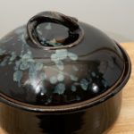 the photograph shows a clay coyote bread baker glazed in midnight garden resting on a small light colored wooden table. the table is in front of a white wall. the bread baker has its lid on. the photograph is lit with white light.