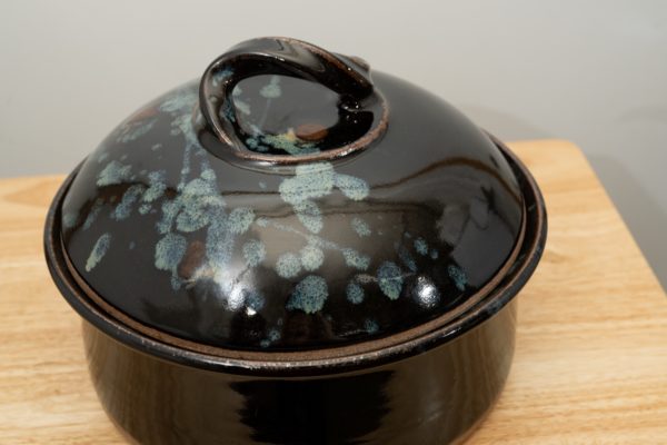 the photograph shows a clay coyote bread baker glazed in midnight garden resting on a small light colored wooden table. the table is in front of a white wall. the bread baker has its lid on. the photograph is lit with white light.