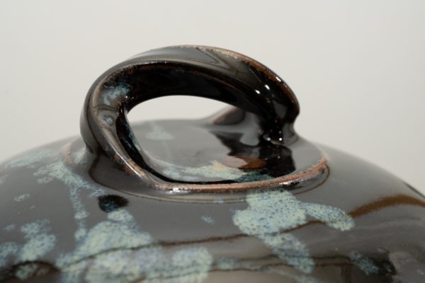 the photograph shows a close up view of the top of a clay coyote bread baker lid. the focus of the photograph is the twisted handle on the top of the break bakers lid. the background is white. the photograph is lit with white light.